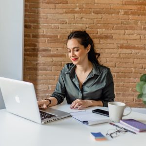 A person works on a laptop at a white table with a coffee cup, notebook, smartphone, eyeglasses, and a potted plant nearby, against a brick wall background.