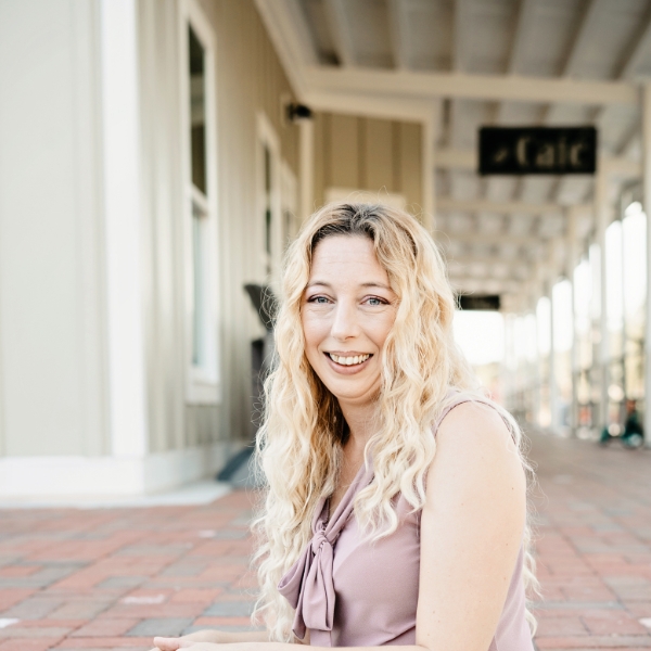 A smiling person with long, wavy blonde hair sits outside near a building with a "cafe" sign, exuding a relaxed and friendly demeanor on a sunny day.