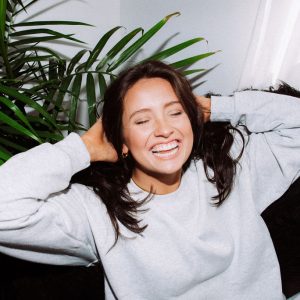 A person is laughing joyfully, hands behind their head, sitting by a window with a green plant and a black cushion, in a well-lit room.