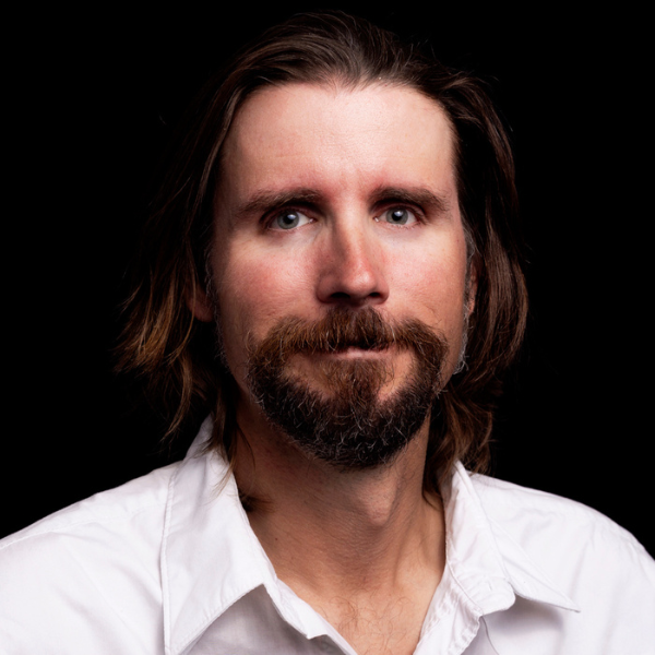 The image shows a person with a medium-length beard and mustache, medium-length brown hair, wearing a white shirt, set against a black background.