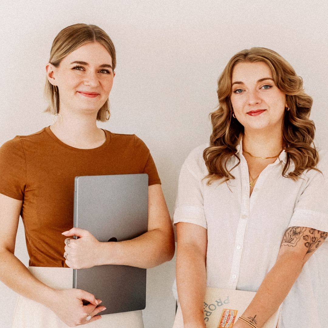 Two smiling people stand side by side against a pale background; one holds a laptop, both dressed in casual work attire, conveying a professional but friendly vibe.