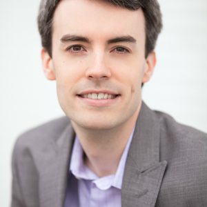 A person with short dark hair, smiling at the camera, wearing a gray blazer over a lavender button-up shirt; neutral background.