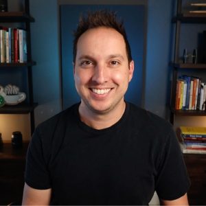 The image shows a smiling person with short hair, wearing a black shirt, in a room with bookshelves and a decorative cup sign in the background.
