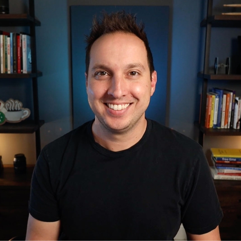 The image shows a smiling person with short hair, wearing a black shirt, in a room with bookshelves and a decorative cup sign in the background.
