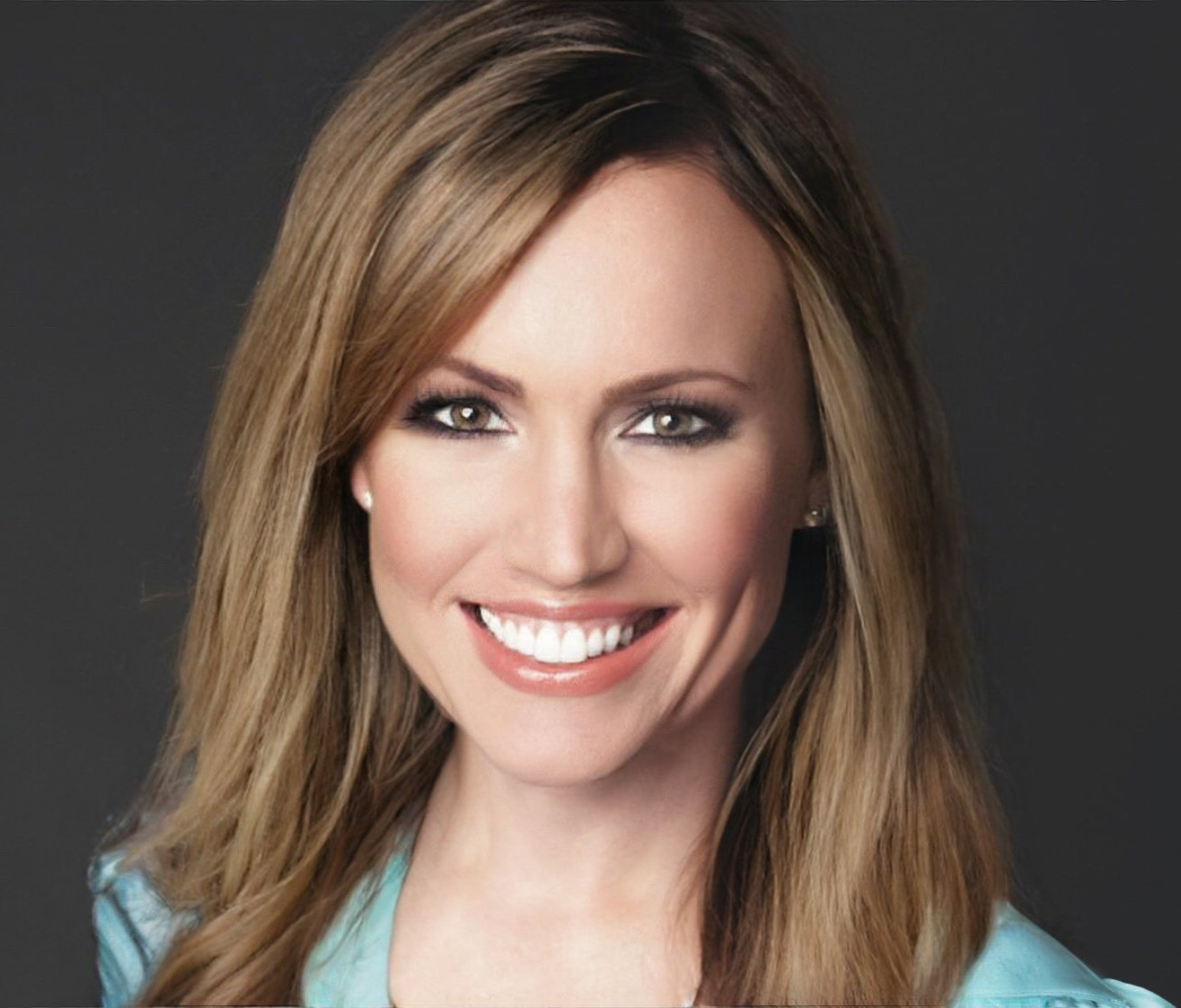 A person with shoulder-length brown hair smiling at the camera. The person wears makeup, earrings, and a turquoise top against a dark background.