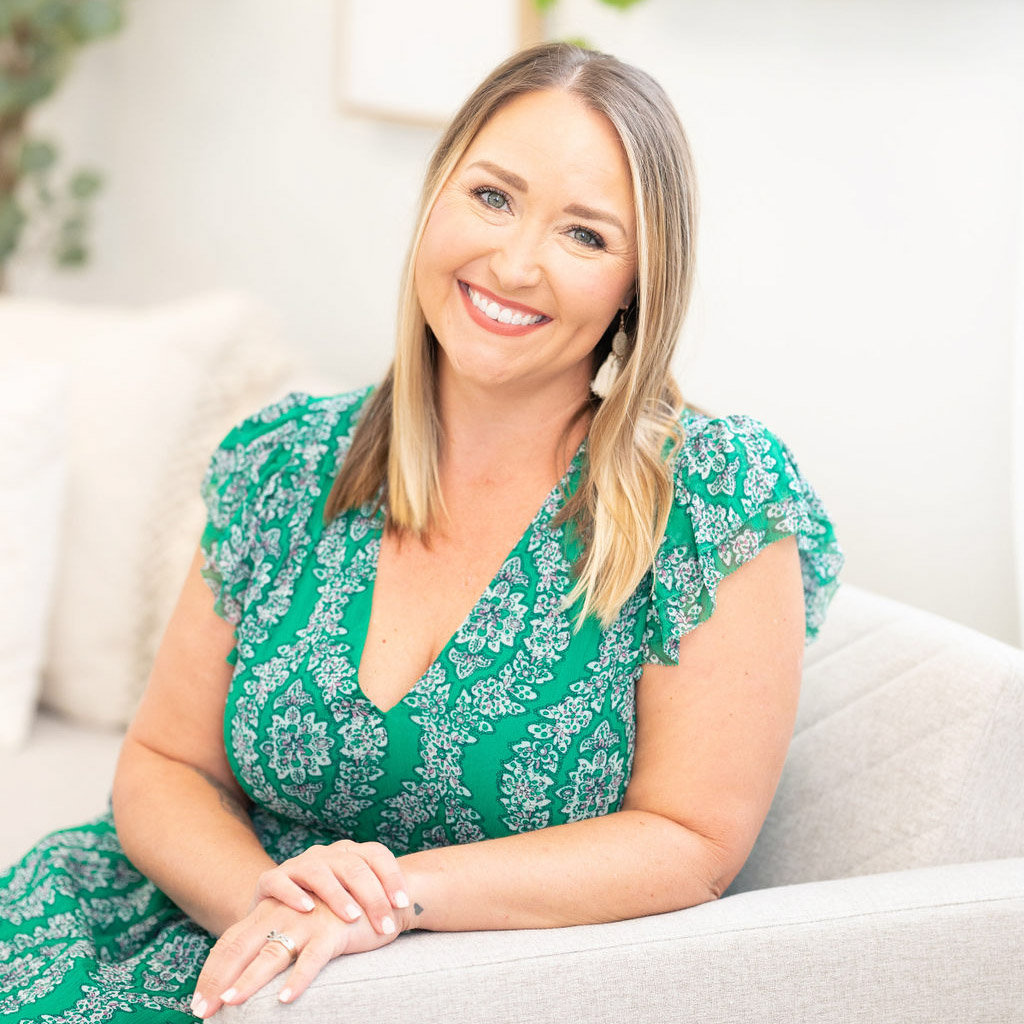 A smiling person sits on a grey sofa, wearing a green patterned dress, in a bright room with a plant and cushions in the background.
