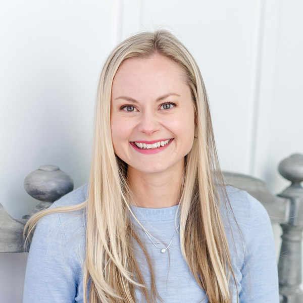 A smiling person with long blonde hair, wearing a light blue top, with a white interior background and decorative objects partially visible.