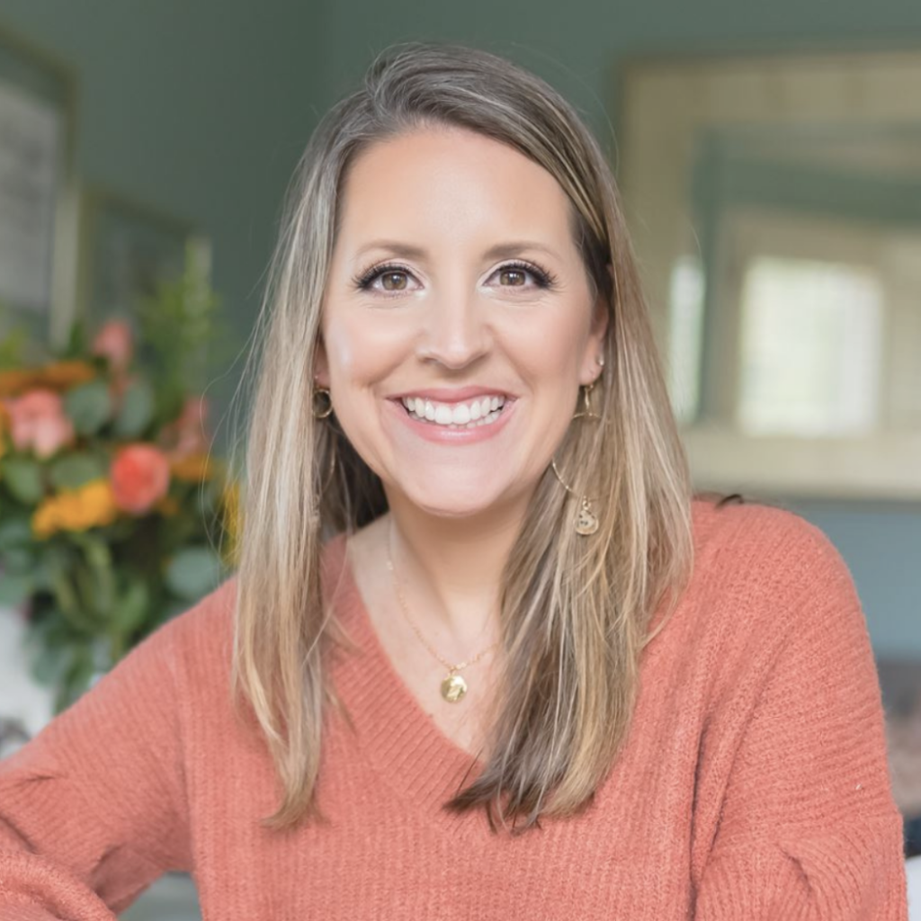 A smiling person with long hair wearing a V-neck sweater poses in a room with green walls and a bouquet of flowers in the background.