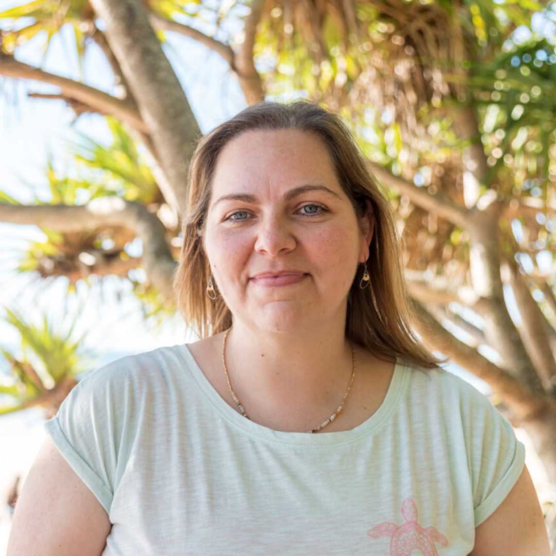 A person is standing outdoors with a natural background featuring trees. They're wearing a light-colored t-shirt with a turtle design and a necklace.