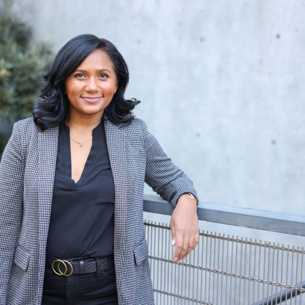 A person is standing outside, leaning against a railing, wearing a houndstooth blazer, black top, and a belt, smiling at the camera.