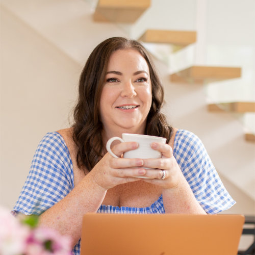 A person sits at a table holding a cup, smiling, with a laptop open. They wear a blue and white gingham top, with a warm indoor background.