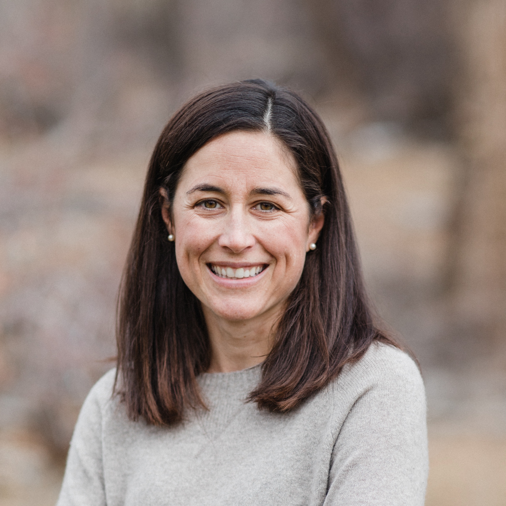 A smiling person with dark hair wearing a gray sweater. Blurred nature background suggesting an informal, outdoor setting. The focus is on the person's face.