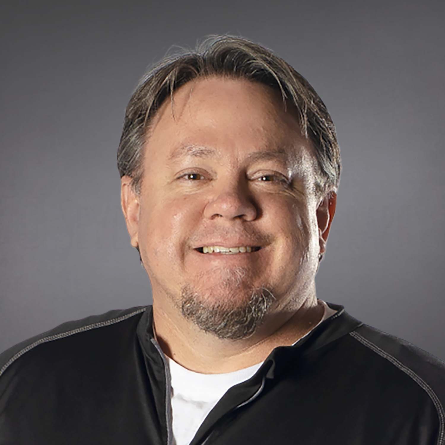The image shows an adult person smiling at the camera, with short brown hair, stubble beard, wearing a black zip-up top, on a grey background.
